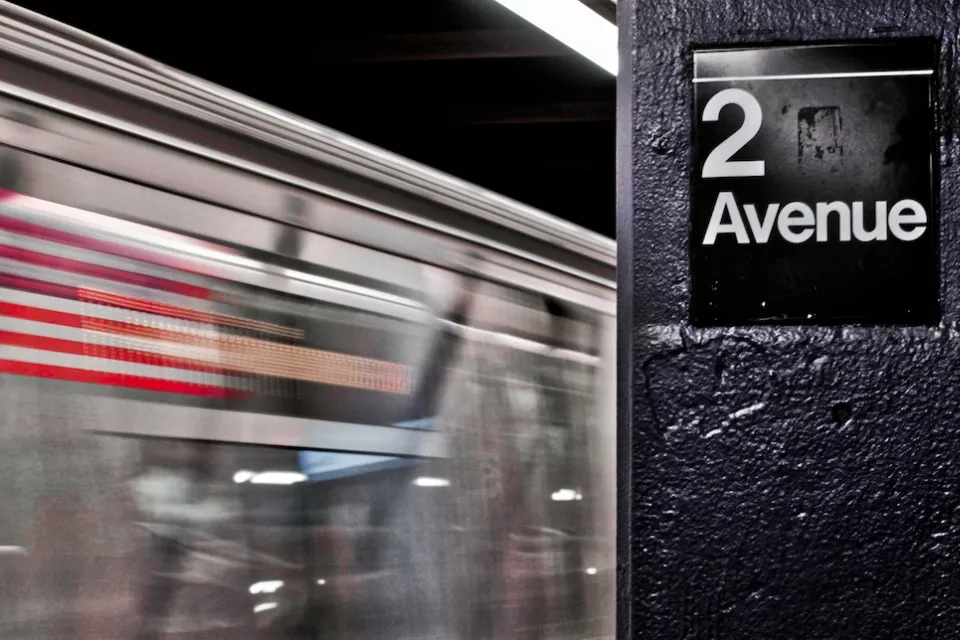 Close-up of 2 Avenue sign on column in NYC subway station with train passing by in background.